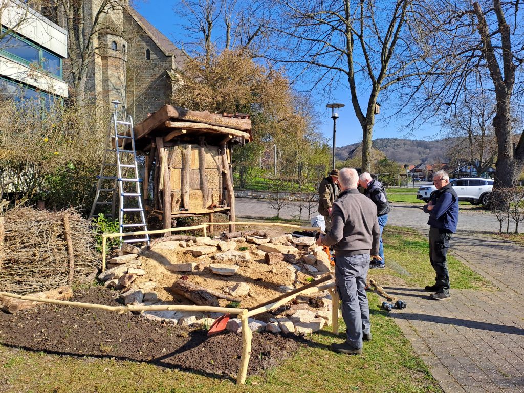 Mehrere Männer betrachten eine im Bau befindliche Steinmauer vor einem Insektenhotel in einem Park bei sonnigem Wetter.