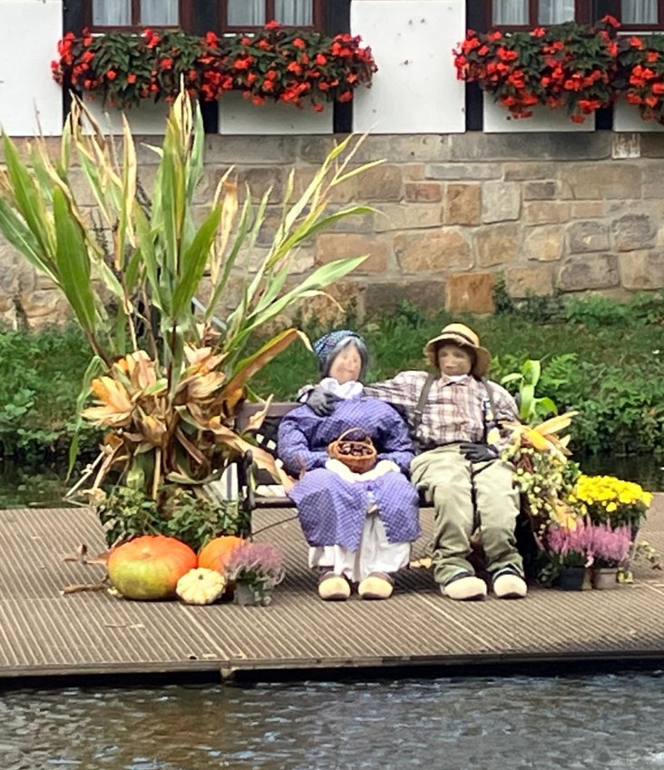 Zwei lebensgroße Figuren in traditioneller Kleidung sitzen auf einer Bank am Wasser, umgeben von Kürbissen, Mais und Blumen.
