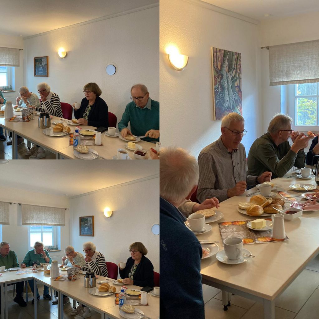 Mehrere Personen sitzen an zwei nebeneinanderstehenden Tischen und frühstücken mit Brötchen, Butter, Aufschnitt, Kaffee und Saft in einem hellen Raum mit weißen Wänden und Fenstern.
