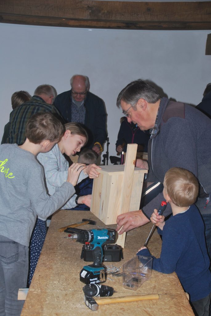 Mehrere Kinder und Erwachsene bauen gemeinsam einen Nistkasten aus Holz an einem langen Tisch mit Werkzeugen wie Akkuschrauber und Hammer.