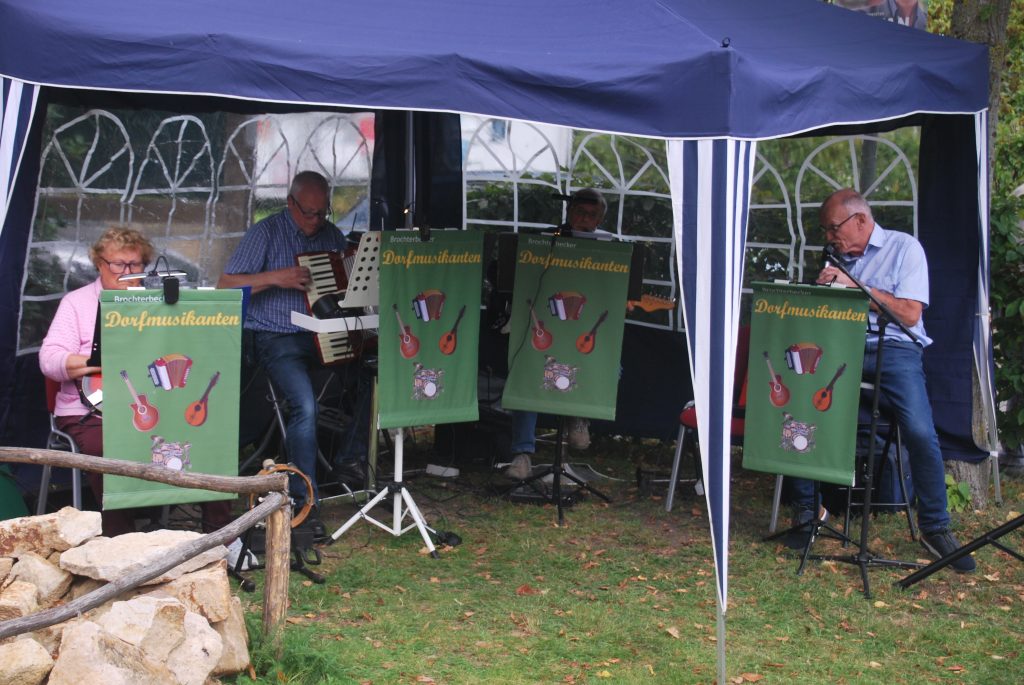 Vier Musiker spielen unter einem blauen Pavillon, vor ihnen hängen grüne Banner mit dem Schriftzug 'Dorfmusikanten' und Instrumentenabbildungen.