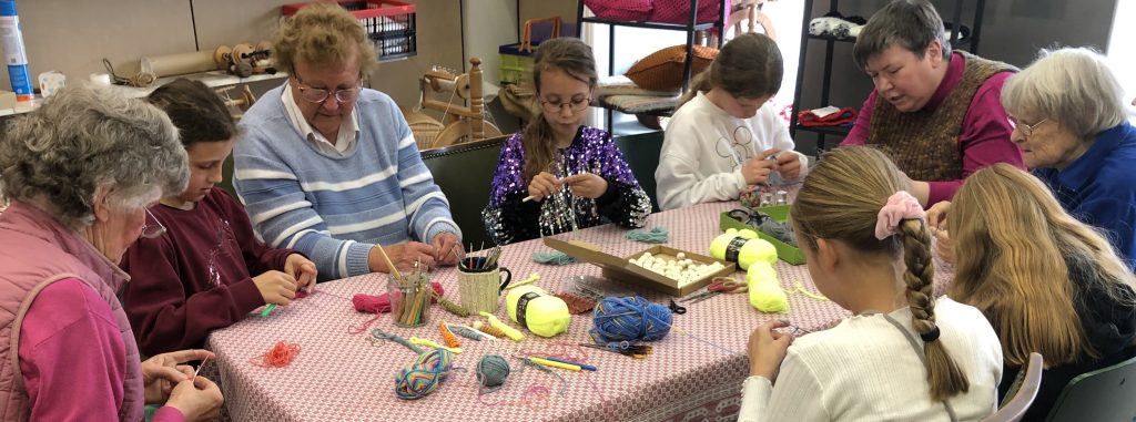 Mehrere Personen sitzen um einen rechteckigen Tisch, der mit einem rot-weiß karierten Tuch bedeckt ist. Auf dem Tisch liegen bunte Wollknäuel, Scheren, Stifte, Pfeifenreiniger, eine Tasse und weitere Bastelmaterialien. Die Personen beschäftigen sich mit Bastelarbeiten und halten verschiedene Materialien in den Händen. Im Hintergrund stehen Holzstühle und ein Regal mit weiteren Gegenständen.