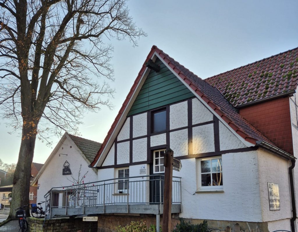 Fachwerkhaus mit weiß verputzten Wänden und dunklen Holzbalken. Das Satteldach ist mit roten Ziegeln gedeckt. Vor dem Gebäude steht ein großer, blattloser Baum. Ein Metallgeländer führt zu einer Rampe am Eingang. Im Hintergrund sind weitere Gebäude mit ähnlicher Architektur zu sehen. Der Himmel ist klar.