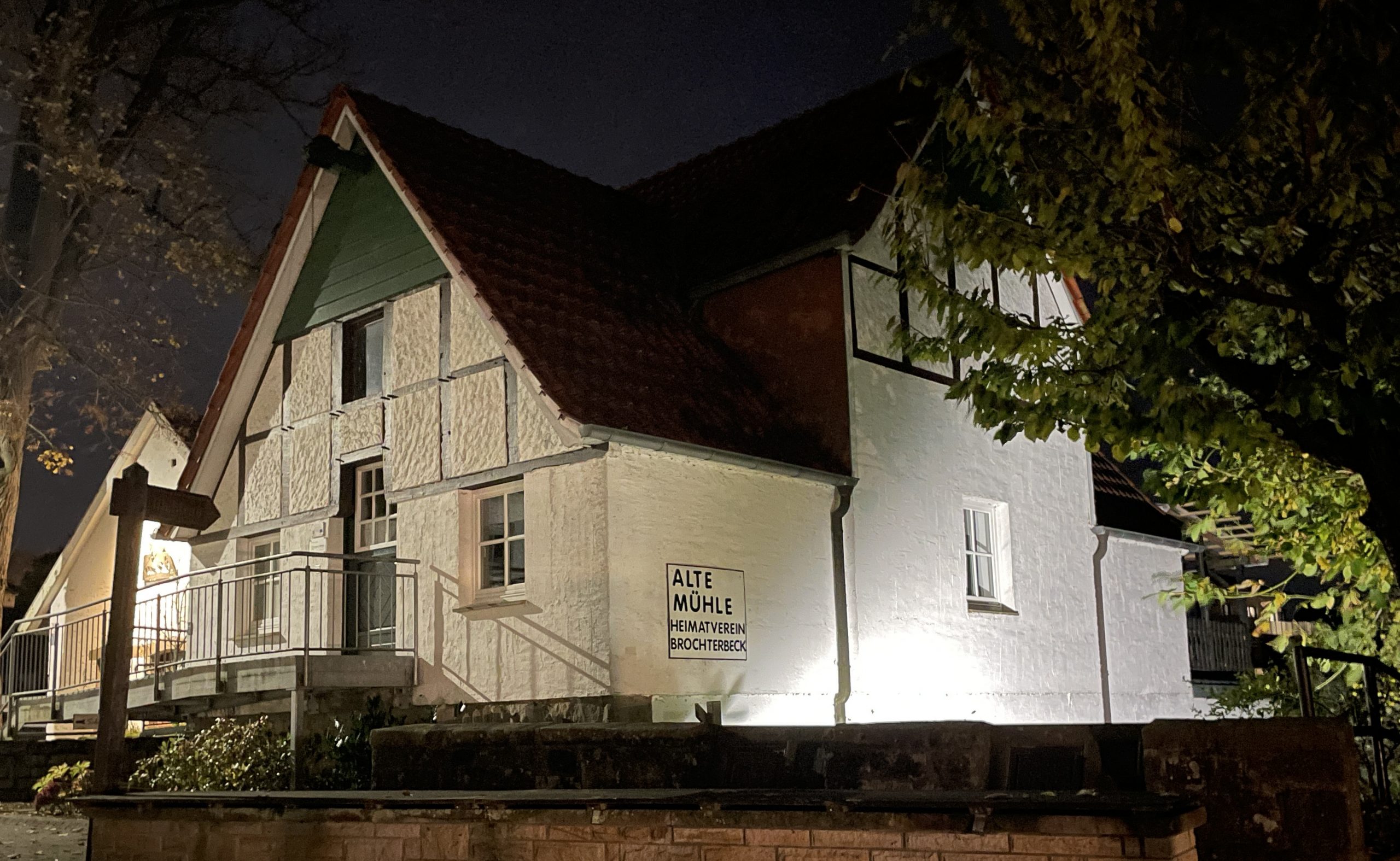 Beleuchtetes Fachwerkhaus mit Schild 'Alte Mühle Heimatverein Brochterbeck' bei Nacht, umgeben von Bäumen.