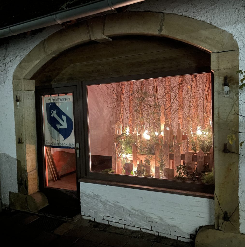 Fensterfront eines alten Gebäudes mit Steinrahmen, dahinter mehrere beleuchtete Kerzen und Tannenzweige als Weihnachtsdekoration.