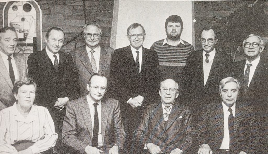 Eine Gruppe von Personen ist in formeller Kleidung vor einer gemauerten Wand und einem weißen Hintergrund versammelt. Die Männer tragen Anzüge und Krawatten, eine Person trägt einen gestreiften Pullover. Eine Frau sitzt vorne links und trägt ein helles Kleid mit auffälligen Falten. Die Aufnahme zeigt den erweiterten Vorstand des Heimatvereins Anfang der 80er Jahre.