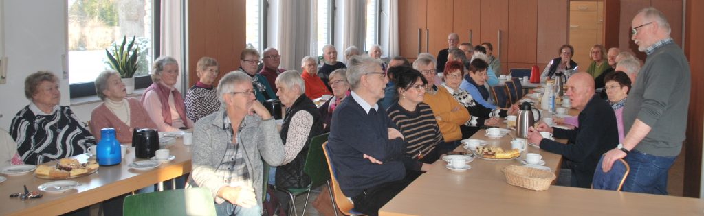 Mehrere Personen sitzen in einem hellen Raum an langen Tischen, auf denen Kaffeetassen, Teller, Kuchen und eine blaue Thermoskanne stehen. Die Fenster lassen viel Tageslicht herein. Die Menschen tragen überwiegend Alltagskleidung wie Pullover und Hemden. Im Hintergrund sind Holzschränke und eine Zimmerpflanze zu sehen. Eine Person steht am rechten Bildrand und spricht zu den Sitzenden.