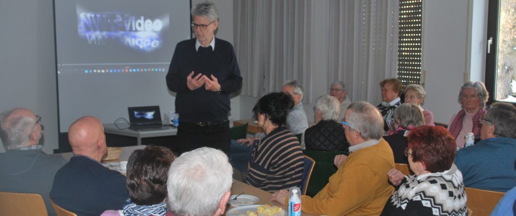 Ein Mann steht vor einer Gruppe älterer Menschen in einem hellen Raum und hält einen Vortrag. Die Zuhörer sitzen an Tischen, auf denen Teller mit Speisen und Getränken stehen. Im Hintergrund ist eine Leinwand mit einer unscharfen Projektion und ein Laptop zu sehen. Die Fenster sind teilweise mit Jalousien abgedeckt, Tageslicht fällt herein. Die Atmosphäre wirkt ruhig und konzentriert.