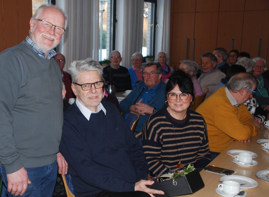 Mehrere Personen sitzen an Tischen in einem hellen Raum mit großen Fenstern und weißen Vorhängen. Auf den Tischen stehen weiße Tassen und Teller, einige mit Speisen belegt. Die Personen tragen überwiegend Freizeitkleidung, darunter Pullover und Hemden. Im Vordergrund ist eine Person mit gestreiftem Oberteil zu sehen. Der Vorsitzende des Heimatvereins Reinhold Engbert bedankt sich bei Walter Tegelmann.