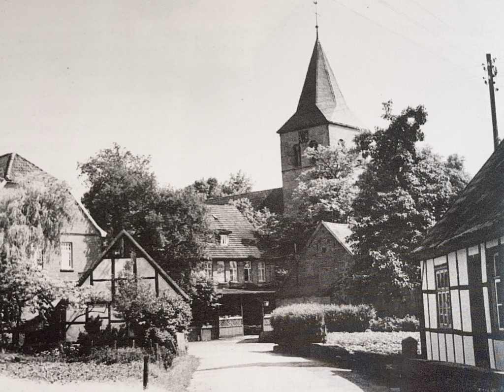 Schwarz-weißes Foto einer ländlichen Szene mit mehreren Fachwerkhäusern und einem Kirchturm im Hintergrund. Die Gebäude sind von Bäumen und Sträuchern umgeben. Die Straße im Vordergrund führt zwischen den Häusern hindurch und ist teilweise von Schatten bedeckt. Die Architektur wirkt traditionell und das Bild vermittelt einen ruhigen, historischen Eindruck. Die Straße läuft auf die Terrasse des Hotels Westfalia zu, das 1973 abgerissen wurde.