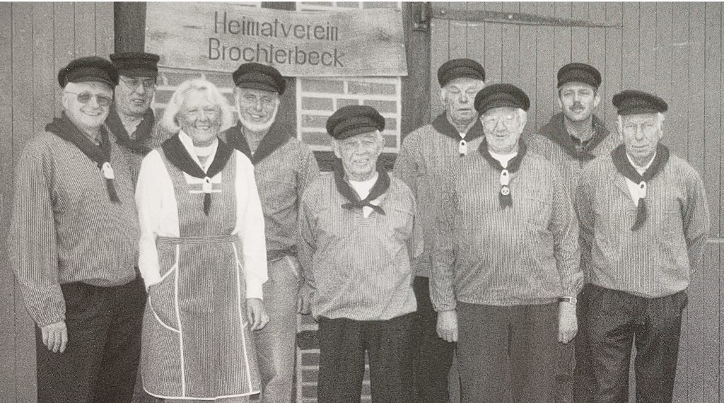 Eine Gruppe von Personen steht nebeneinander vor einer Holzwand mit einem Schild, auf dem 'Heimatverein Brochterbeck' steht. Die Personen tragen einheitliche Kleidung mit dunklen Mützen, Halstüchern und Pullovern. Eine Person trägt eine helle Bluse und eine Schürze. Die Aufnahme ist schwarz-weiß und zeigt den erweiterten Vorstand des Brochterbecker Heimatvereins 1999.