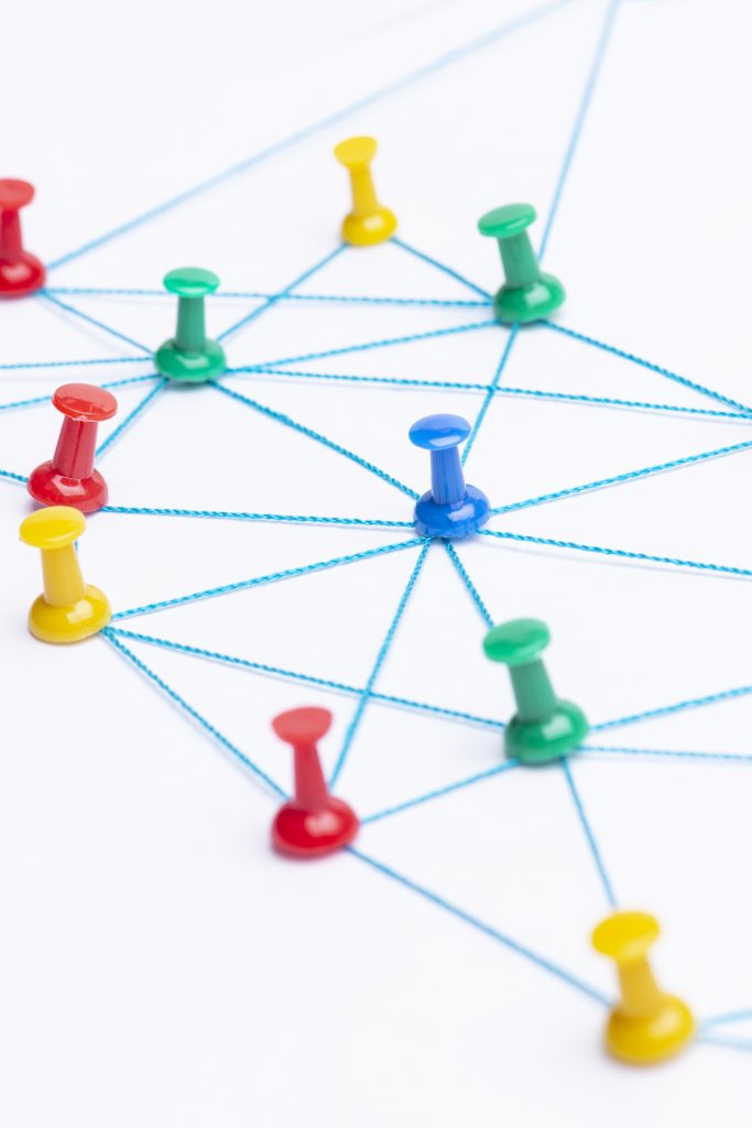 Mehrfarbige Stecknadeln sind auf einer weiรen Flรคche angeordnet und durch blaue Fรคden miteinander verbunden. Die Fรคden verlaufen in verschiedenen Richtungen und bilden ein Netzwerk aus Linien und Knotenpunkten. Die Stecknadeln sind in den Farben Rot, Gelb, Grรผn und Blau gehalten.