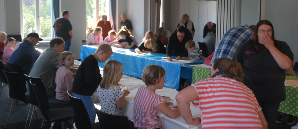 Mehrere Kinder und Erwachsene sitzen und stehen an langen, mit bunten Tischdecken bedeckten Tischen in einem hellen Raum. Die Kinder beschäftigen sich mit Bastel- oder Malaktivitäten, während einige Erwachsene danebenstehen oder sich unterhalten. Im Hintergrund sind große Fenster mit Tageslicht und weitere Personen zu sehen. Die Atmosphäre wirkt lebhaft und gemeinschaftlich.