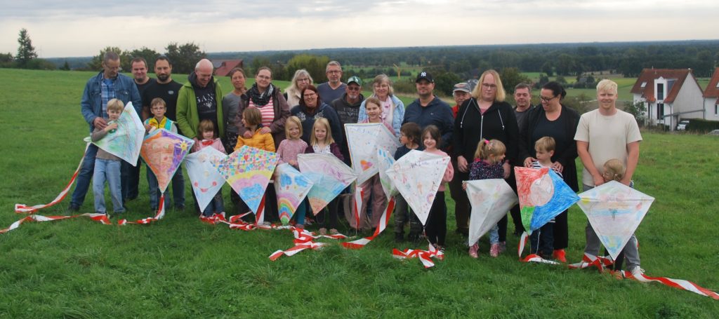 Eine Gruppe von Erwachsenen und Kindern steht auf einer Wiese. Die Kinder halten bunte Drachen mit langen, rot-weißen Bändern. Im Hintergrund sind Häuser und eine weite Landschaft mit Feldern und Bäumen zu sehen. Der Himmel ist bewölkt.